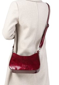 Femme portant un manteau beige et portant un sac à bandoulière en cuir laqué rouge foncé avec une sangle ajustable sur le dos.