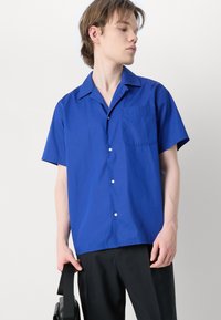 Camisa de botão de manga curta em azul brilhante, com um bolso no peito, colarinho plano e textura suave. Usada com calças pretas e uma bolsa.