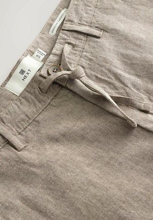 Smėlio spalvos austi kelnės su įprasta siaurėjančia forma. Yra rudas mygtuko užsegimas, diržo kilpos ir etiketė, nurodanti dydį 34.