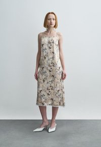 Robe nuisette en soie beige à motif floral, avec fines bretelles et bordure en dentelle à l'ourlet. Le mannequin porte des escarpins blancs à bout pointu sur un sol en béton.