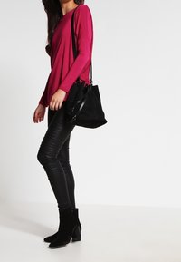 Langärmliges fuchsiefarbenes Oberteil, schwarze Leggings aus Kunstleder, schwarze Absatz-Stiefeletten und eine schwarze Wildleder-Handtasche mit verstellbarem Tragegurt.
