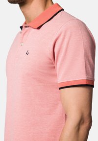 Różowa koszulka polo z teksturowanej tkaniny, kontrastowy kołnierz i mankiety w kolorze pomarańczowym z czarnym wykończeniem, z dwoma guzikami i logo na klatce piersiowej.