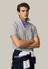 Giovane con polo grigio chiaro con dettaglio e stemma blu sul colletto, maglione blu navy e bianco legato in vita, braccia conserte.