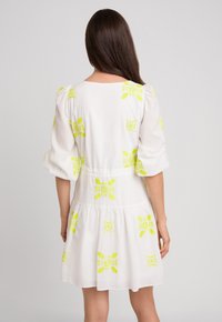 Biała sukienka z wyrazistym żółtym haftem kwiatowym, bufiastymi rękawami, dopasowanym w pasie i warstwowym fasonem spódnicy. Gładka tekstura materiału.