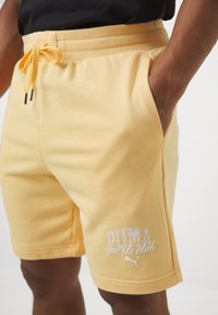 Žute kratke hlače od mekanog materijala s elastičnim pojasom, detaljem na vezanje i bijelim logotipom "PUMA sports club" sa strane.