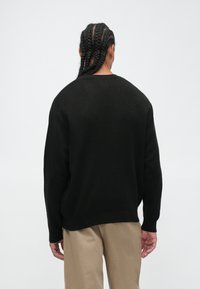 Personne avec de longues tresses portant un pull noir et un pantalon beige, se tenant de dos contre un fond blanc uni.