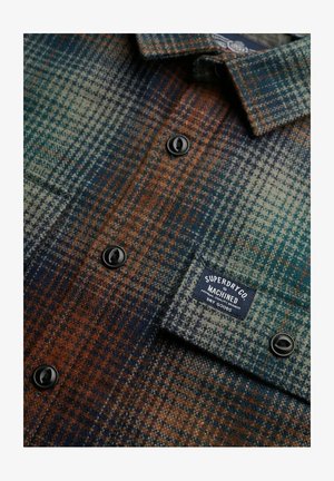 Camicia di flanella a quadri nei toni del verde, blu e marrone. Presenta bottoni neri, un'etichetta a patch e un materiale texture simile alla lana.