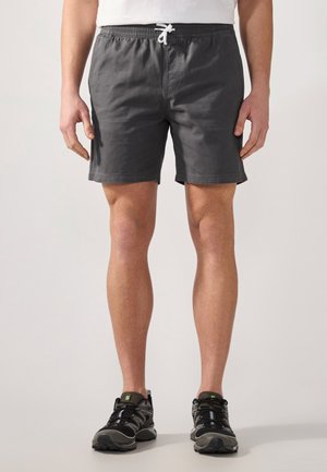 Mies, joka on pukeutunut tummanharmaisiin narutaskushortseihin ja mustaharmaisiin juoksukenkään, seisoo yksinkertaista taustaa vasten.