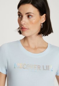 Jasnoniebieska bawełniana koszulka z okrągłym dekoltem i krótkimi rękawami, z napisem "ODKRYJ ŻYCIE" w iryzującym nadruku na przodzie.