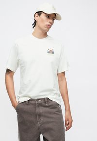 Valge lühikeste varrukatega T-särk, mille vasakul rinnal on mägimaastiku graafiline trükk. Sobitatud hallide, lõdva lõikega pükstega.