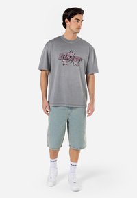 Grå kortermet t-skjorte med grafisk design som viser stjerner og tekst. Brukt med lyseblå denimshorts og hvite joggesko.