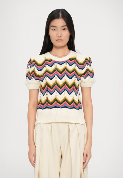 Giovane donna con lunghi capelli scuri che indossa un top lavorato a maglia crema con motivi zigzag colorati e maniche corte a sbuffo, abbinato a pantaloni beige chiaro.