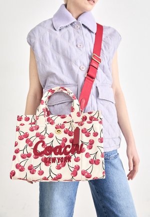 Personne portant une veste matelassée sans manches lilas clair et un jean tient un sac fourre-tout blanc avec un imprimé cerises rouges et le texte "Coach New York".