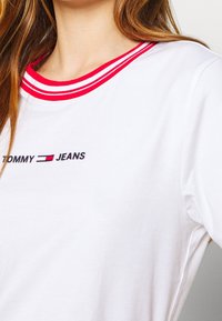 Camiseta de algodón blanca con escote redondo que presenta ribeteado de rayas rojas y blancas; logo "TOMMY JEANS" impreso en negro en el pecho.