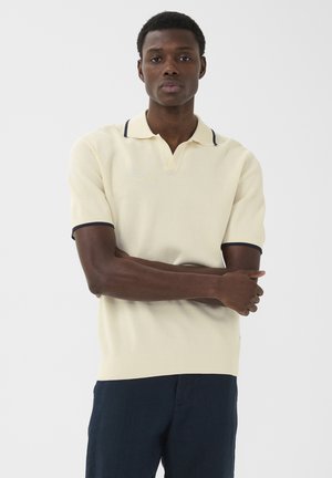 Junger Mann mit kurzen Haaren, der ein cremefarbenes Kurzarm-Poloshirt mit dunklem Rand und dunklen Hosen trägt, steht mit verschränkten Armen vor weißem Hintergrund.