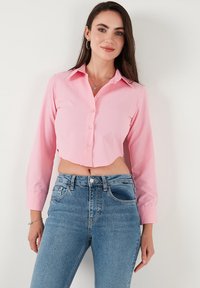 Pink cropped button-down shirt with long sleeves, featuring a classic collar and smooth texture, paired with high-waisted blue denim jeans.