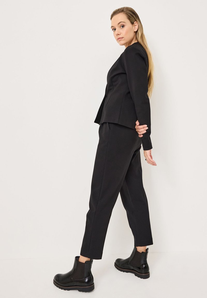 Traje negro a medida con un blazer entallado y pantalones ajustados, complementado con botines negros brillantes, sobre un fondo neutro.