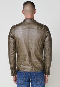 Uomo che indossa una giacca di pelle marrone e jeans blu, in piedi rivolto all'indietro con le mani rilassate ai lati su uno sfondo semplice.