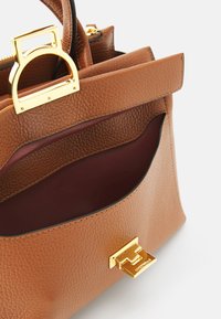 Sac à main en cuir marron avec une surface texturée, quincaillerie dorée et une poche intérieure bordeaux foncé. Il présente une poignée supérieure distincte et une fermeture.