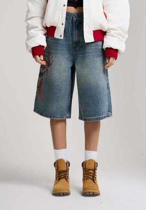 Person trägt weite Jeansshorts mit besticktem Design, weiße Socken, beige Schnürstiefel, eine kurz geschnittene Jacke mit roten Bündchen, steht vor einem einfarbigen Hintergrund.