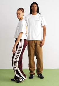 Weiße T-Shirts, eines mit Grafik, kombiniert mit maroon- und cremefarbenen gestreiften Jogginghosen und braunen Cargo-Hosen. Schwarze Sneakers mit Streifen.
