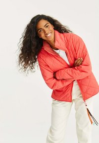 Kurtka w kolorze koralowym o pikowanej fakturze, z wysokim kołnierzem, przednim zamkiem błyskawicznym i dekoracyjnym sznurkiem na dole. Noszona na białej bluzce, w zestawieniu z jasnymi spodniami.