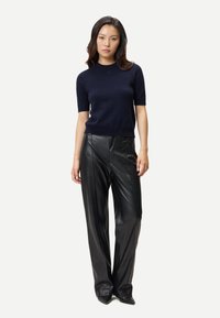 Kvinna klädd i en marinblå kortärmad tröja och svarta, höga skidläderbyxor med vida ben, stående mot en enkel ljus bakgrund.