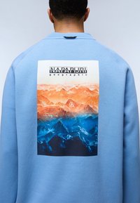 Felpa azzurra con una grande grafica di una montagna in arancione e blu sul retro, con il logo "Napapijri" in alto.