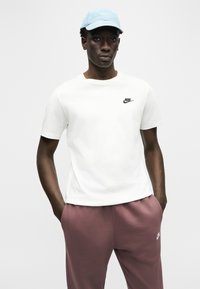 Młody mężczyzna w jasnoniebieskiej czapce Nike, białej koszulce Nike i fioletowych spodniach dresowych Nike, stojący z rękami w kieszeniach na jednolitym tle.