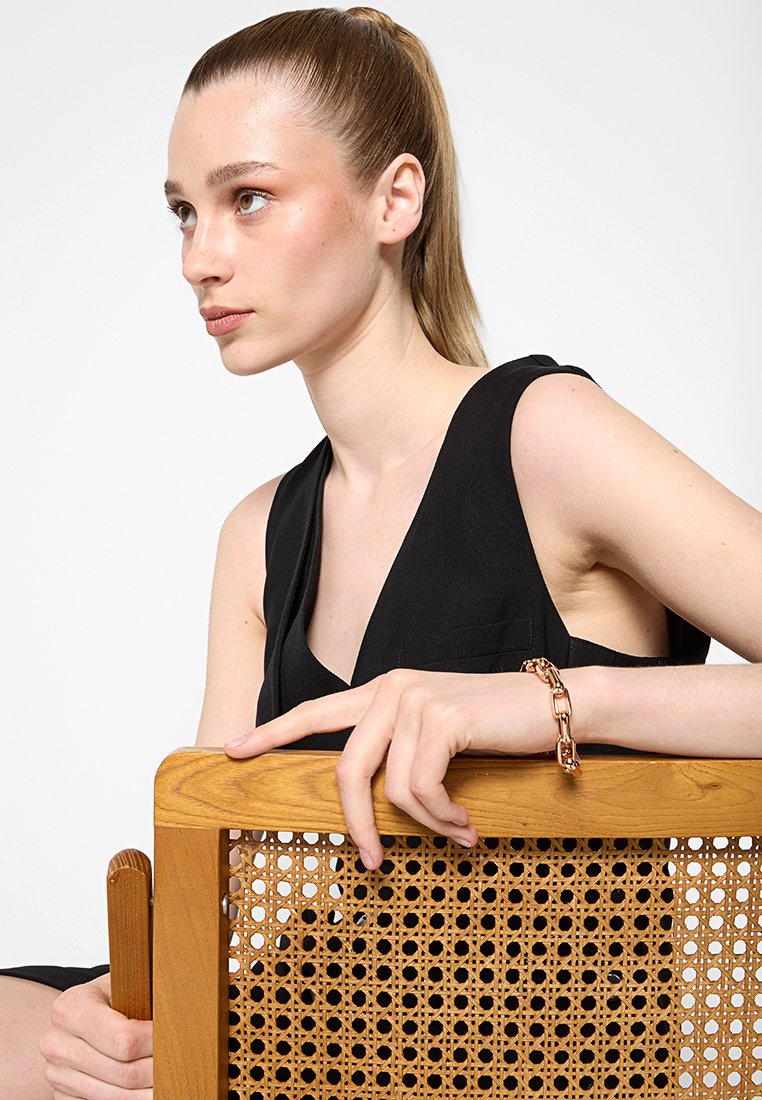 Junge Frau mit Pferdeschwanz, die ein schwarzes ärmelloses Top trägt, sitzt hinter einem Holzstuhl mit geflochtenem Rücken und trägt ein goldenes Kettenarmband.