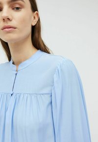 Svetlomodrá blúzka s okrúhlym výstrihom, skladanými detailami a dlhými rukávmi. Mäkšia tkanina, gombíkové akcenty na golieri a hladká textúra.