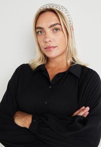 Black button-up shirt with long, gathered sleeves. Model wears a beaded headband. Smooth fabric texture, minimalistic design. Light background.