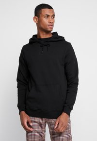 Felpa nera con cappuccio regolabile, tasca frontale e polsini a costine. Design semplice, realizzata in tessuto morbido, indossata con pantaloni a quadri.