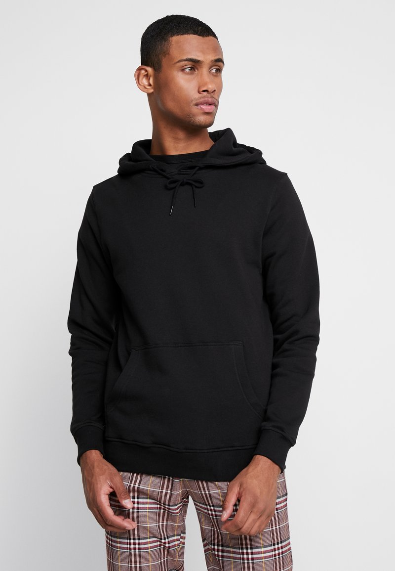 Felpa nera con cappuccio regolabile, tasca frontale e polsini a costine. Design semplice, realizzata in tessuto morbido, indossata con pantaloni a quadri.