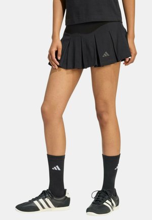 Donna con gonna nera plissettata, calzini neri Adidas e sneakers nere Adidas con strisce bianche, in piedi davanti a uno sfondo uniforme.