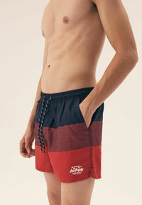 Szorty kąpielowe męskie z granatowym i czerwonym paskiem, elastyczną talią ze sznurkiem oraz bocznymi kieszeniami. Logo nadrukowane na dolnej części przodu.