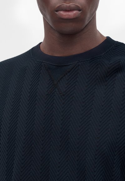 Gros plan sur la partie inférieure du visage et le cou d'une personne portant un pull bleu foncé texturé avec un motif chevron subtil.