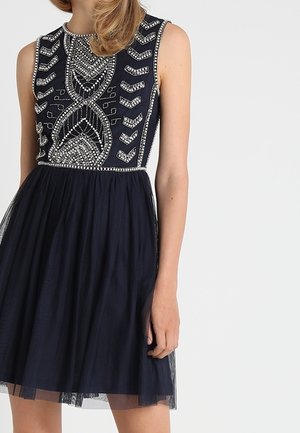 Mouwloze marineblauwe jurk met zilveren kralenversieringen op het lijfje en een doorschijnende gelaagde rok, gedragen door een persoon met een lichte huidtint.