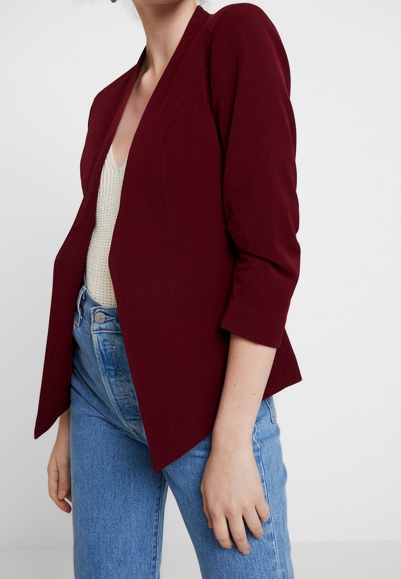 Personne portant un blazer bordeaux à manches trois-quarts par-dessus un top en maille blanc et un jean bleu taille haute sur un fond uni.