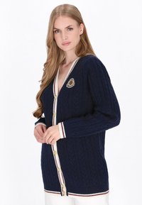 Laivaston sininen cable-knit neuletakki, jossa on ribatut kaulus ja hihansuut, kultasävyiset napit, valkoiset ja burgundinväriset yksityiskohdat sekä brodattu emblem.