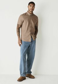 Kortærmet, button-down skjorte i lysebrun stof, parret med løse blå jeans og brune ruskindsko. Simpelt design; ingen mønstre.