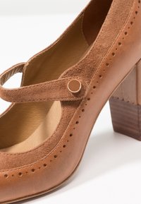 Chaussure Mary Jane en cuir marron avec des détails en daim, bride à bouton rond, motif perforé et un talon bloc solide sur fond blanc.