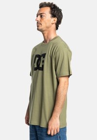 Camiseta de algodón verde oliva con cuello redondo y mangas cortas, que presenta un gran logotipo negro en la parte frontal y un corte informal.