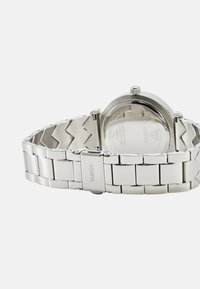 Uhani iz nerjavečega jekla s poliranim zaključkom, zapestnica z medsebojnimi povezavami in okroglo ohišje. Na hrbtni strani je prikazan logotip blagovne znamke in podrobnosti.