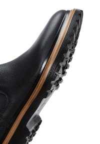 Zapato de cuero negro con superficie texturizada, que cuenta con una suela de goma negra con tacos de tracción y un acento marrón a lo largo del borde.