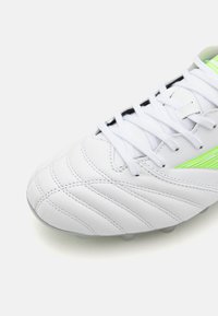 Scarpa sportiva bianca con tomaia in pelle texturizzata, dettagli in verde e suola grigia con tacchetti sagomati per una migliore aderenza.