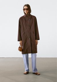 Brun, knälång kappa med knäppning, sidofickor och hög krage. Bärs med ljusblå jeans och platta sandaler.