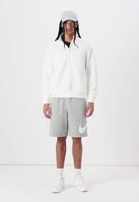 Maglione bianco in pile con zip a quarto, abbinato a pantaloni corti grigi con un grande logo Nike bianco. Indossato con un cappellino grigio chiaro e sneaker bianche.