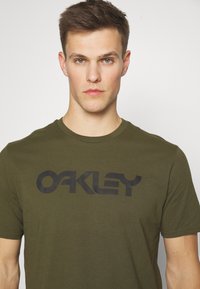 Oliven grøn T-shirt i bomuld med et stort sort "OAKLEY" logo henover brystet. Klassisk rund hals og korte ærmer.