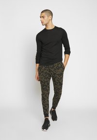 T-shirt noir à manches longues associé à un pantalon de jogging camouflage vert, avec une coupe ajustée et des poignets côtelés, complété par des chaussures de sport noires.